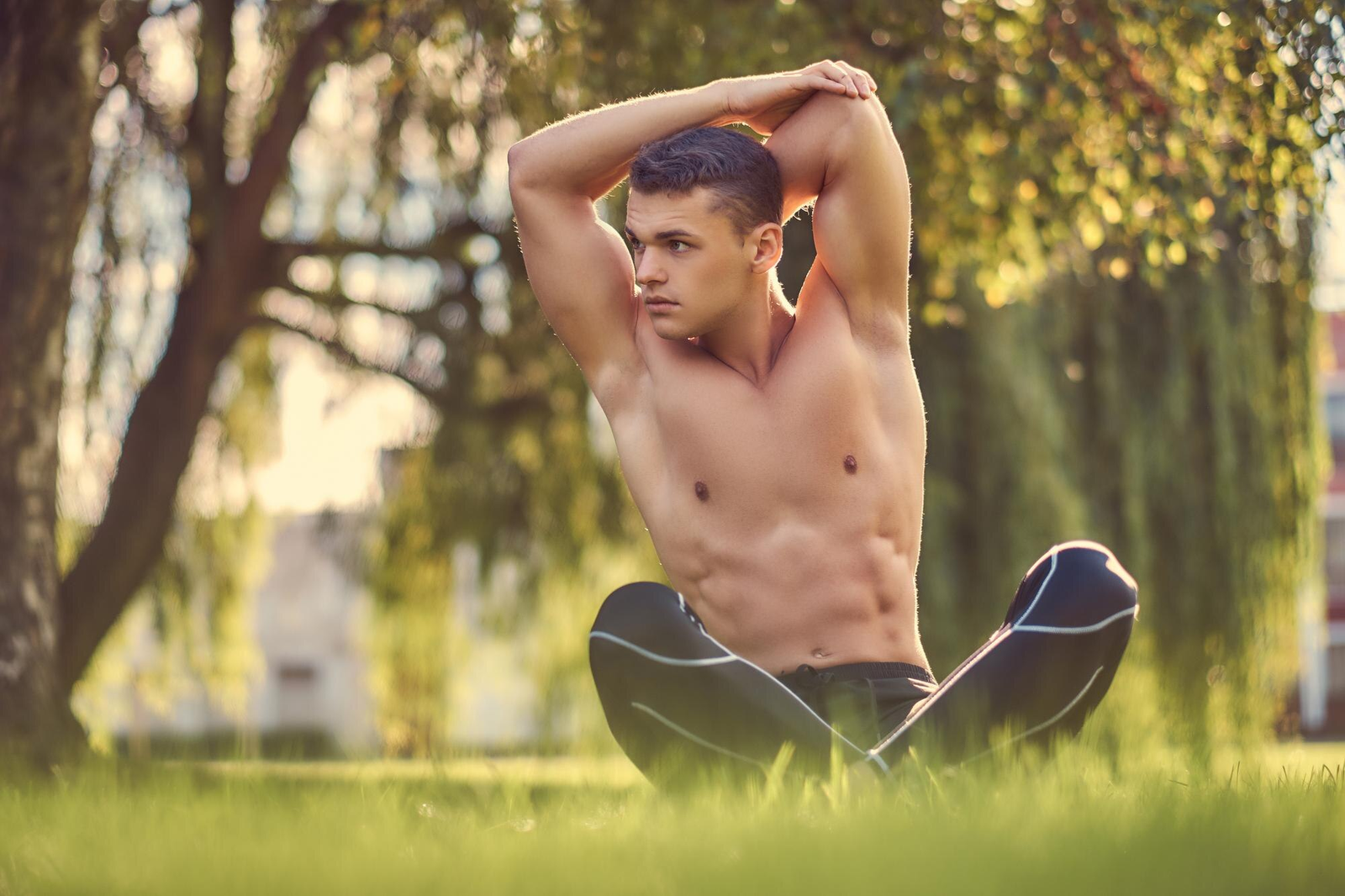 Homem jovem musculoso sentado na grama ao ar livre, alongando os braços - melhor horário para praticar yoga